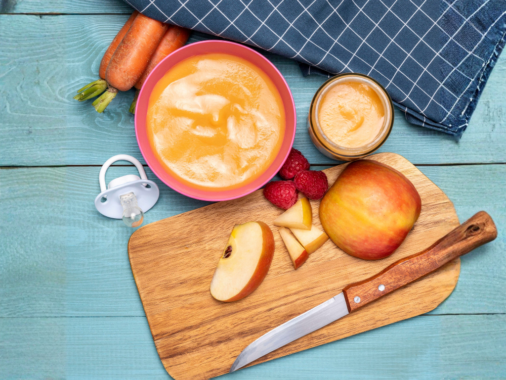 Mesa con una tabla de madera que contiene una manzana roja, rodajas de manzana, un cuchillo, un tazón de puré de frutas, un frasco de puré de manzana y unas frambuesas, con una mantelería a cuadros y zanahorias al fondo.